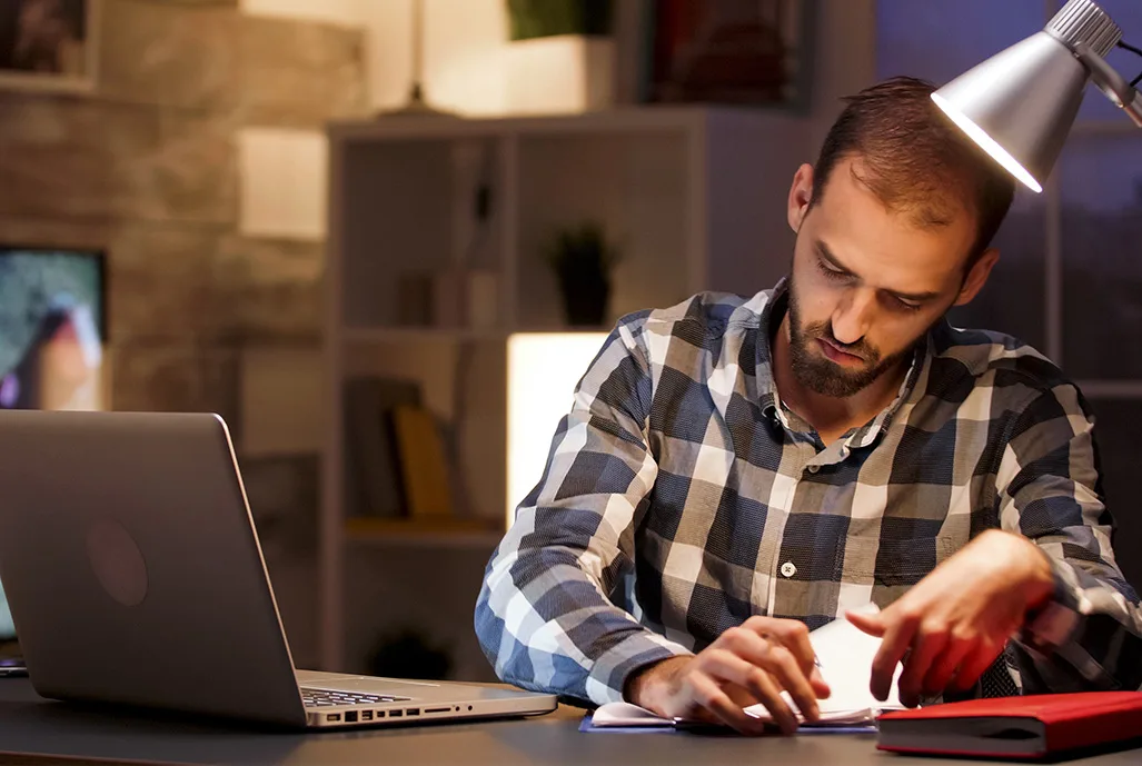 Empreendedor escrevendo anotações à mesa de trabalho à noite, ao lado de um notebook, sob a luz de uma luminária
