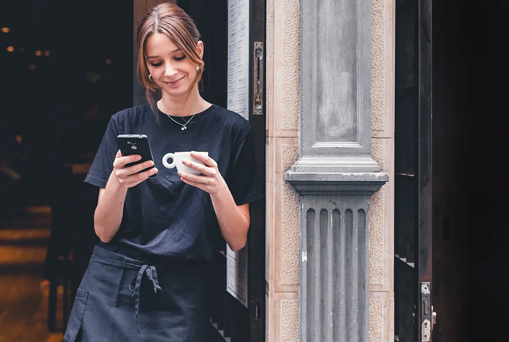 Atendente de cafeteria sorrindo enquanto olha o celular na porta do estabelecimento, com uma xícara de café na mão